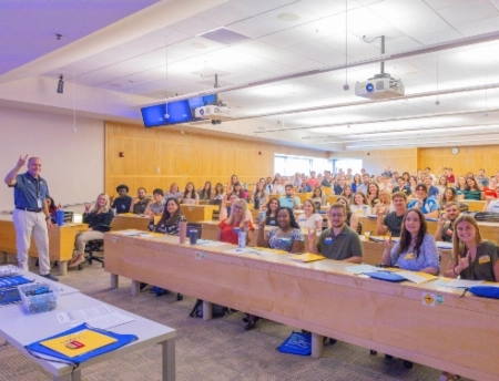 Dean Melchert and a new group of pharmacy students make the Roo hand sign in a large School of Pharmacy lecture hall.